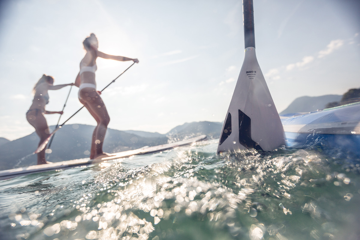Zwei Personen beim Stand-Up-Paddling auf einem See, im Hintergrund Berge